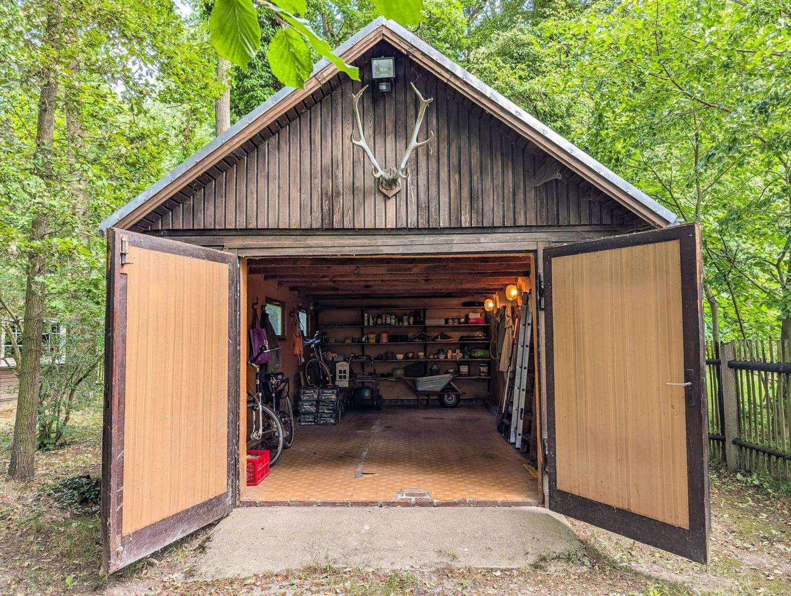 Wooden shed with antlers mounted on the peak, doors wide open to a cluttered workshop with shelves, tools, a bicycle, and a ladder inside.