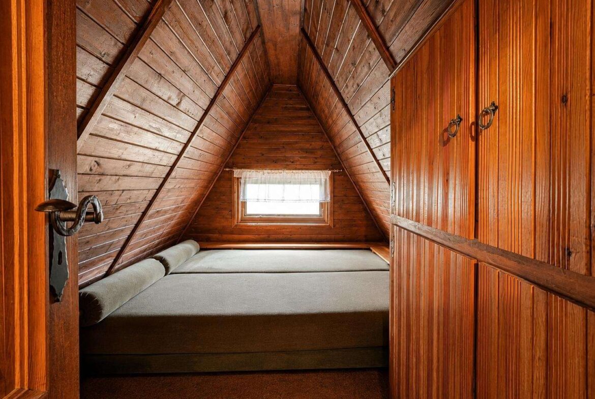 Cozy wooden attic bedroom with paneled walls, a small window at the far end, and a bed along the floor under a pitched ceiling.