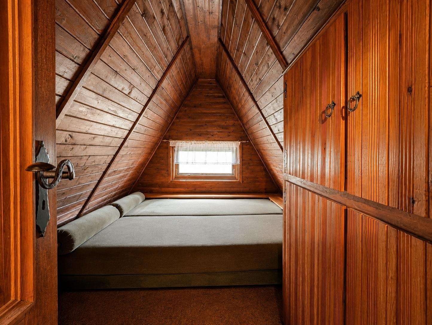 Cozy wooden attic bedroom with paneled walls, a small window at the far end, and a bed along the floor under a pitched ceiling.