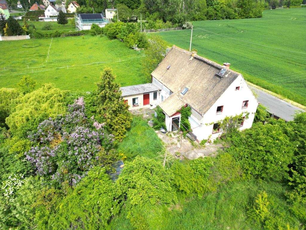 White two-story house with a sloped roof amid lush green fields and trees, a small outbuilding nearby and a country road on the side.