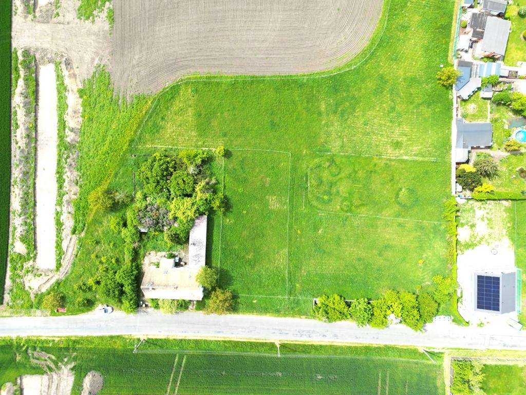 Aerial view of a rural property: green fields, a small house with trees, a road below, and surrounding gardens/plots with another building rooftop solar panels on the right.