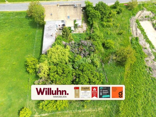 Aerial view of a house surrounded by trees and green fields, with a large Willuhn Immobilien sign in the foreground.