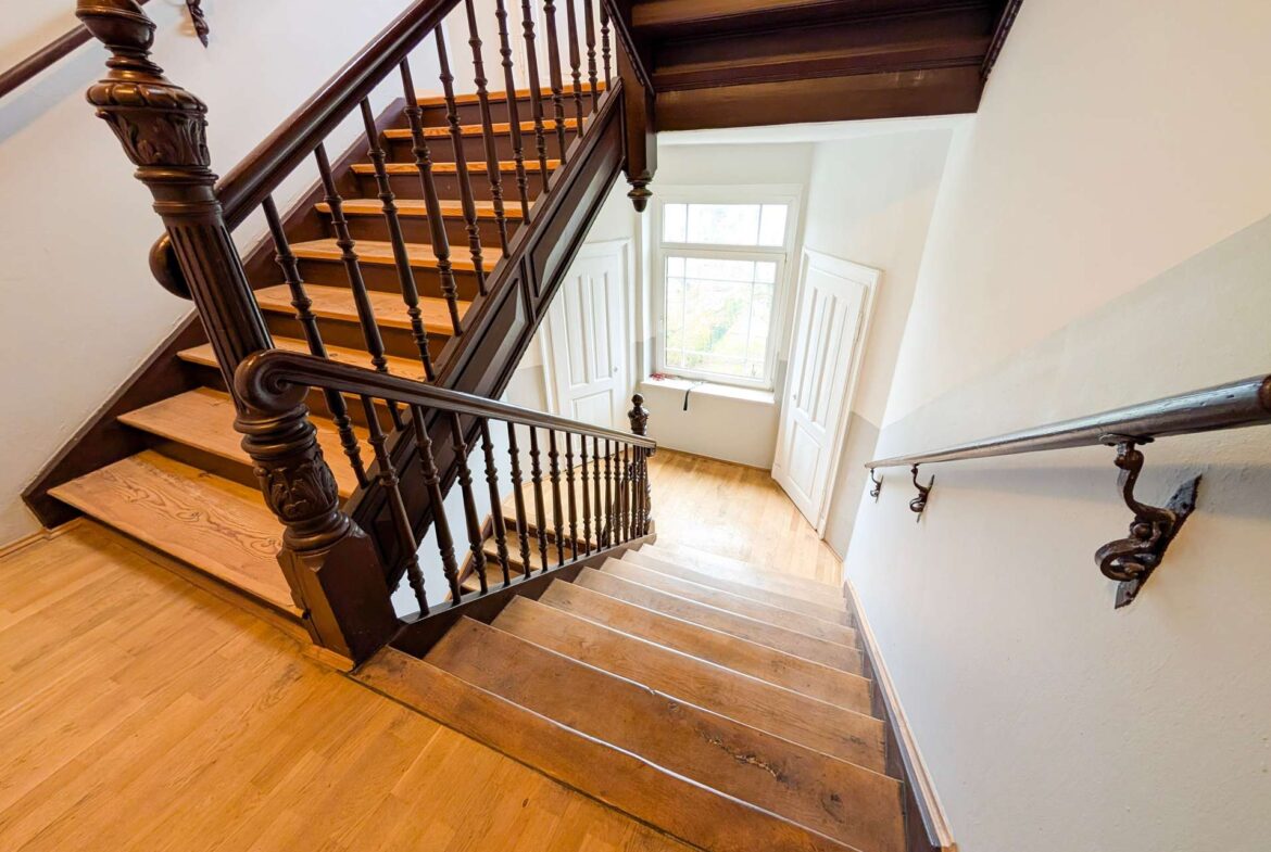 Dark wood staircase with carved newel post and spindles, light wooden steps, white walls, and a window at the landing below.
