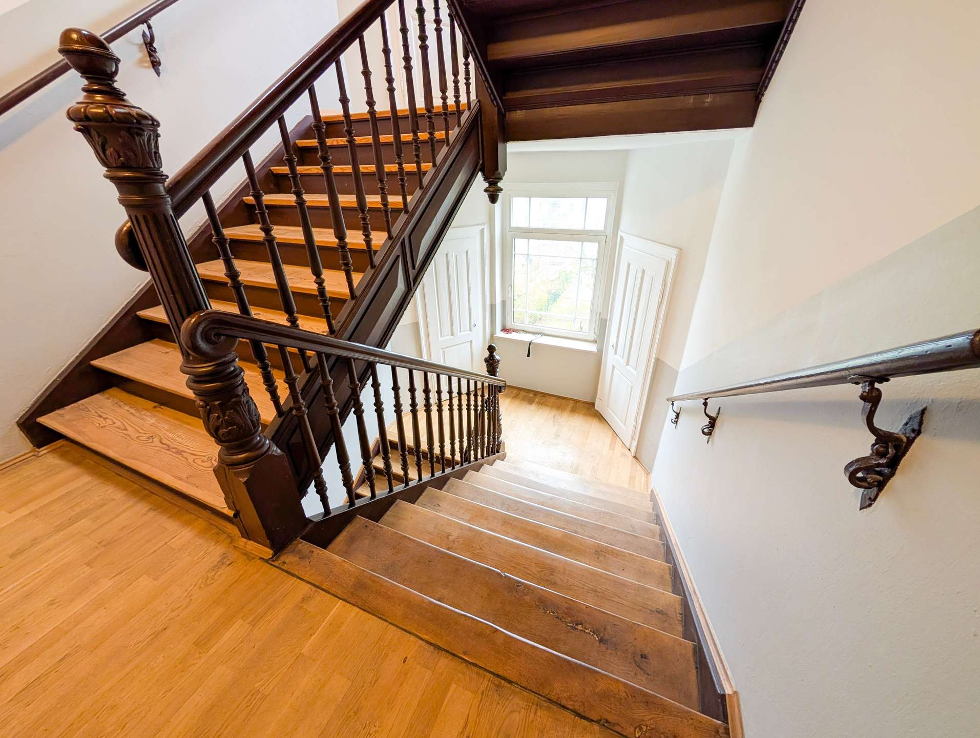 Dark wood staircase with carved newel post and spindles, light wooden steps, white walls, and a window at the landing below.
