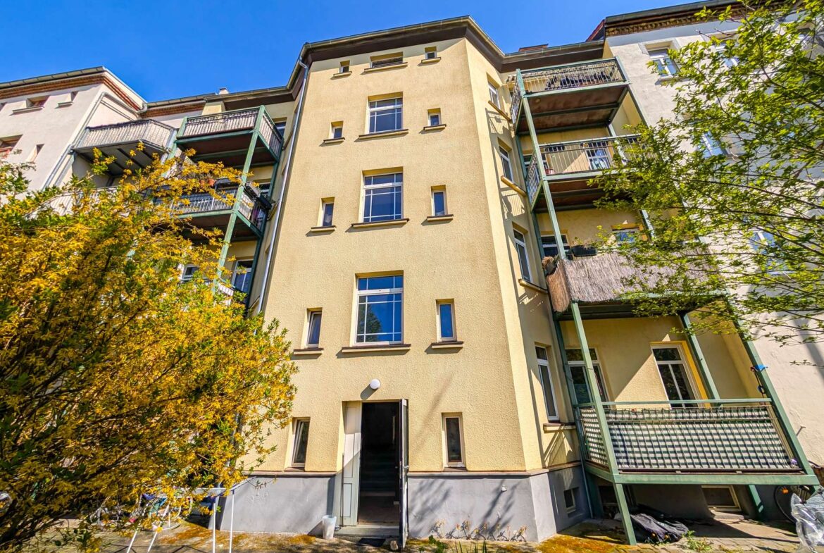 Yellow apartment building with green balconies and an open entry, foreground of autumn-leaved trees against a bright blue sky.