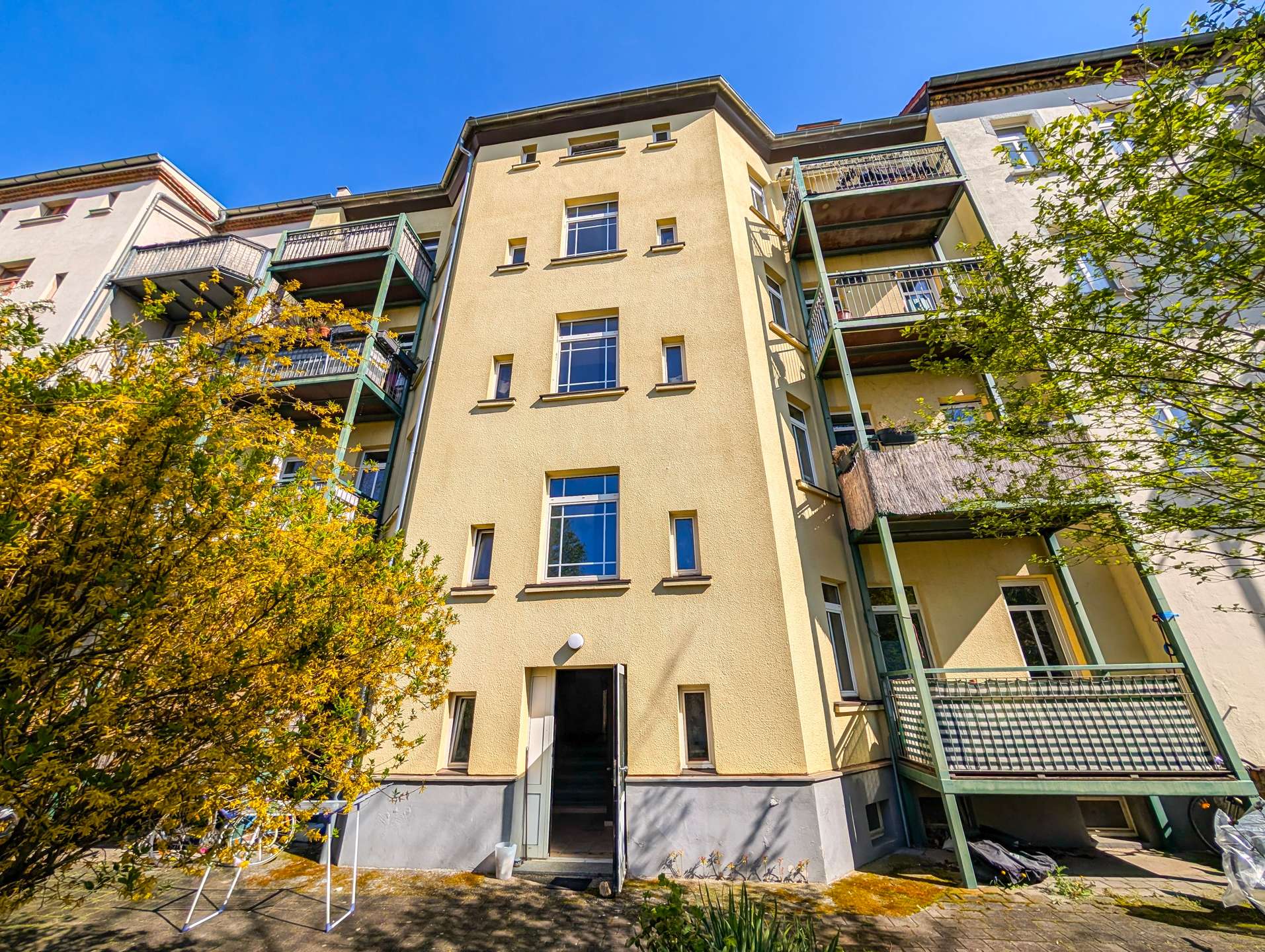 Yellow apartment building with green balconies and an open entry, foreground of autumn-leaved trees against a bright blue sky.
