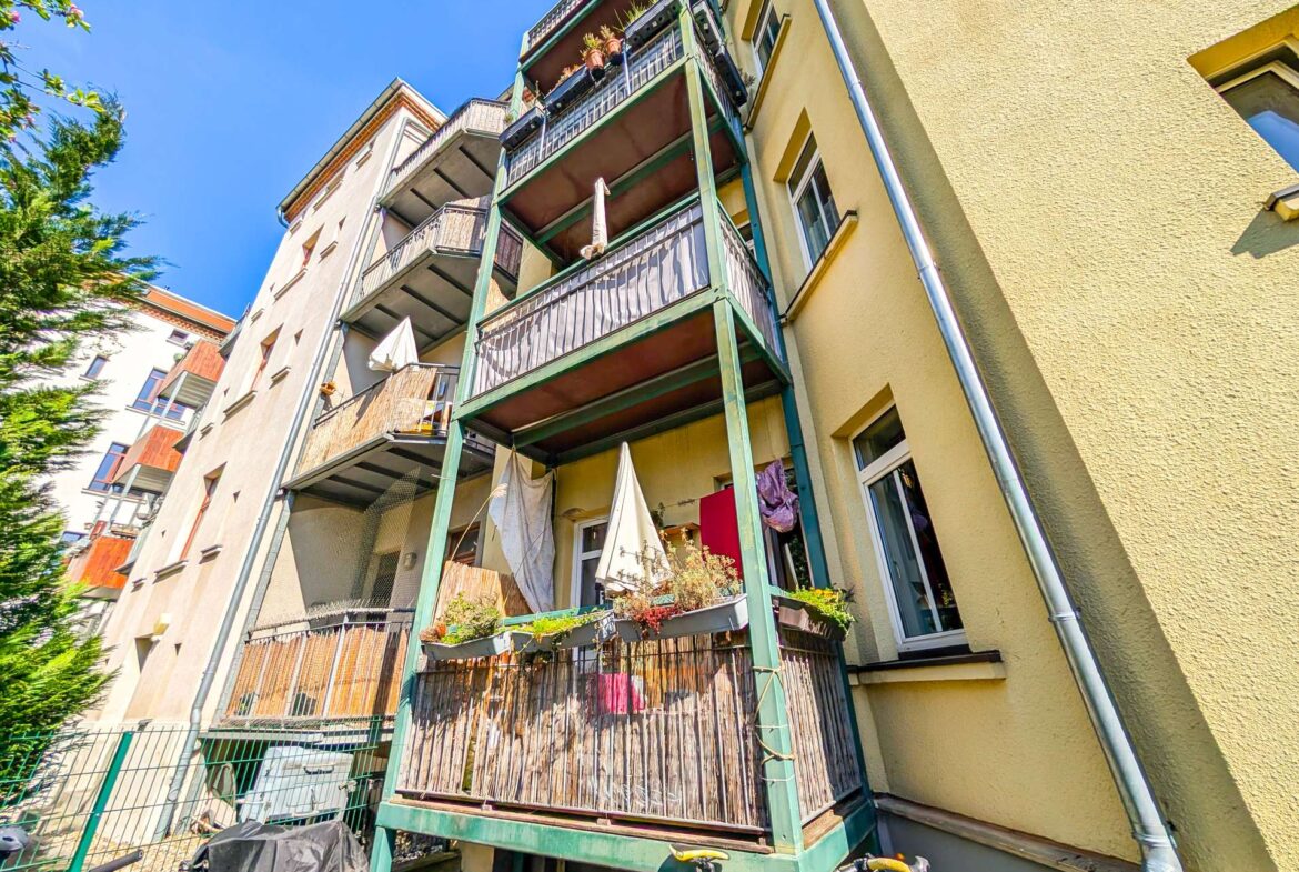 Ground-level view of a multi-story apartment building with green-framed balconies, potted plants, and umbrellas against a bright blue sky, with bicycles in the foreground.