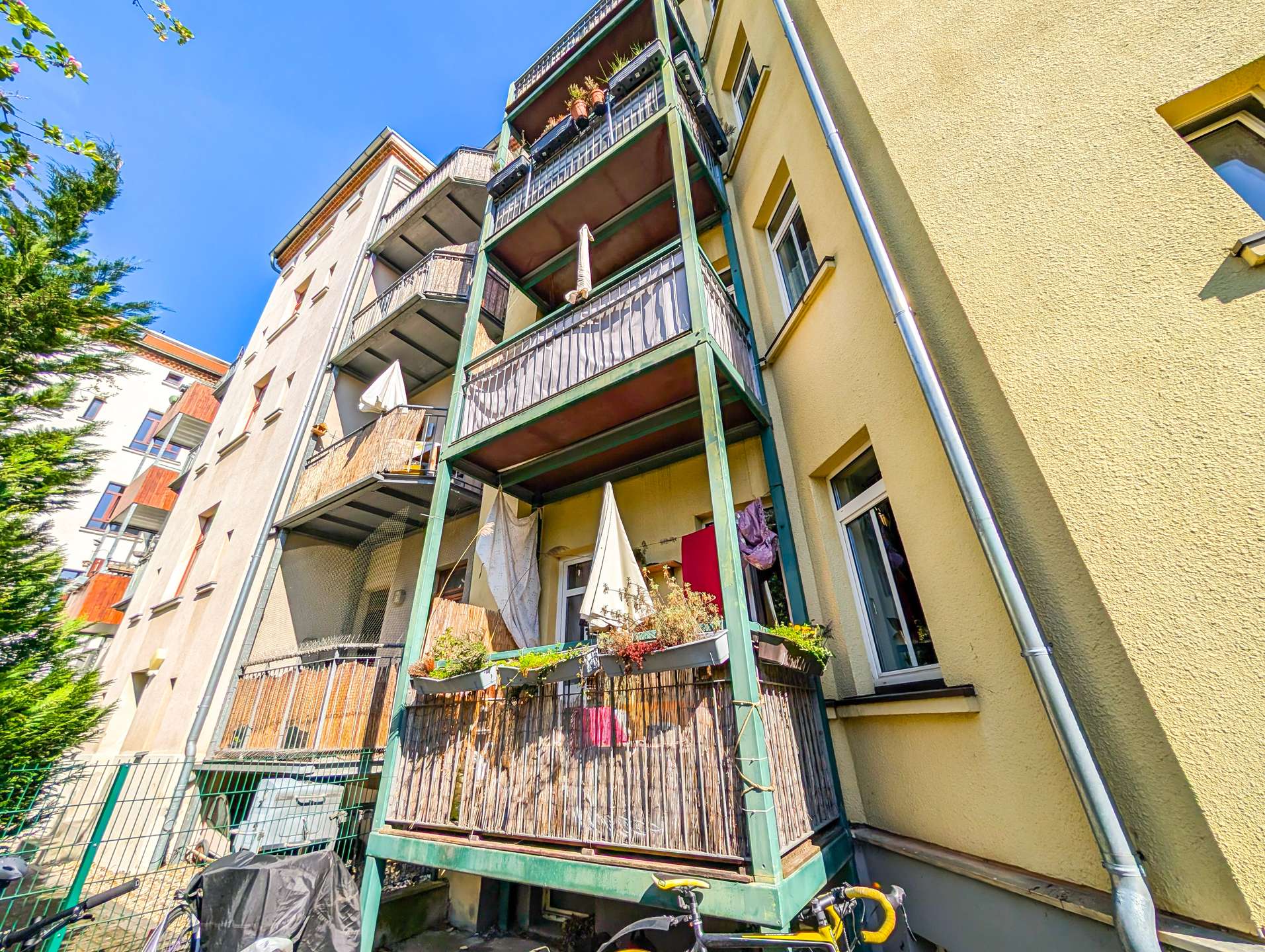 Ground-level view of a multi-story apartment building with green-framed balconies, potted plants, and umbrellas against a bright blue sky, with bicycles in the foreground.