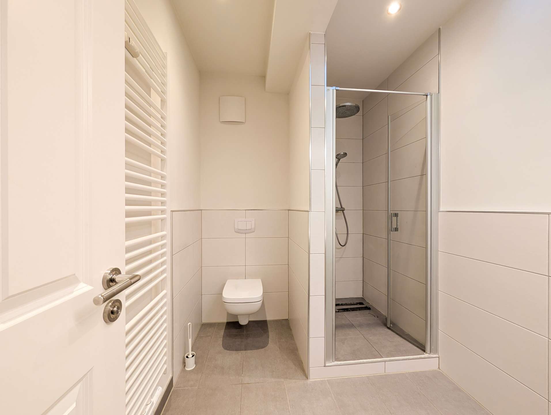 Small modern bathroom with a wall-mounted toilet, white tile walls, and a glass shower enclosure on the right; a vertical white towel radiator is on the left.