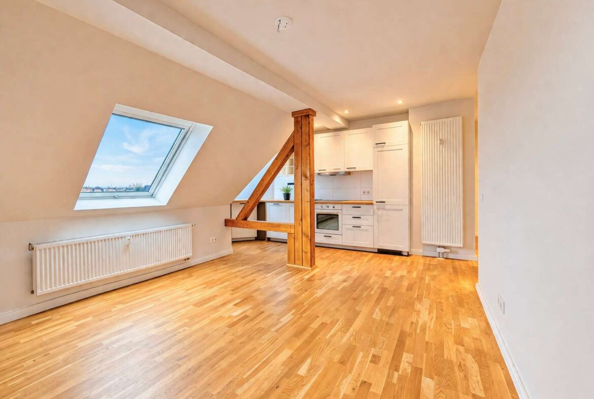 Bright open-plan attic space with sloped walls, a large skylight, a wooden support beam, and a white kitchen at the rear.