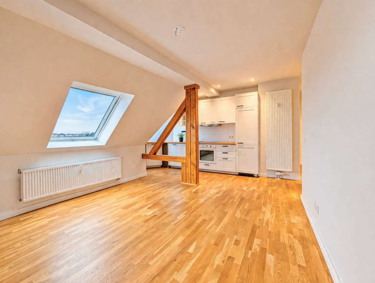 Bright open-plan attic space with sloped walls, a large skylight, a wooden support beam, and a white kitchen at the rear.