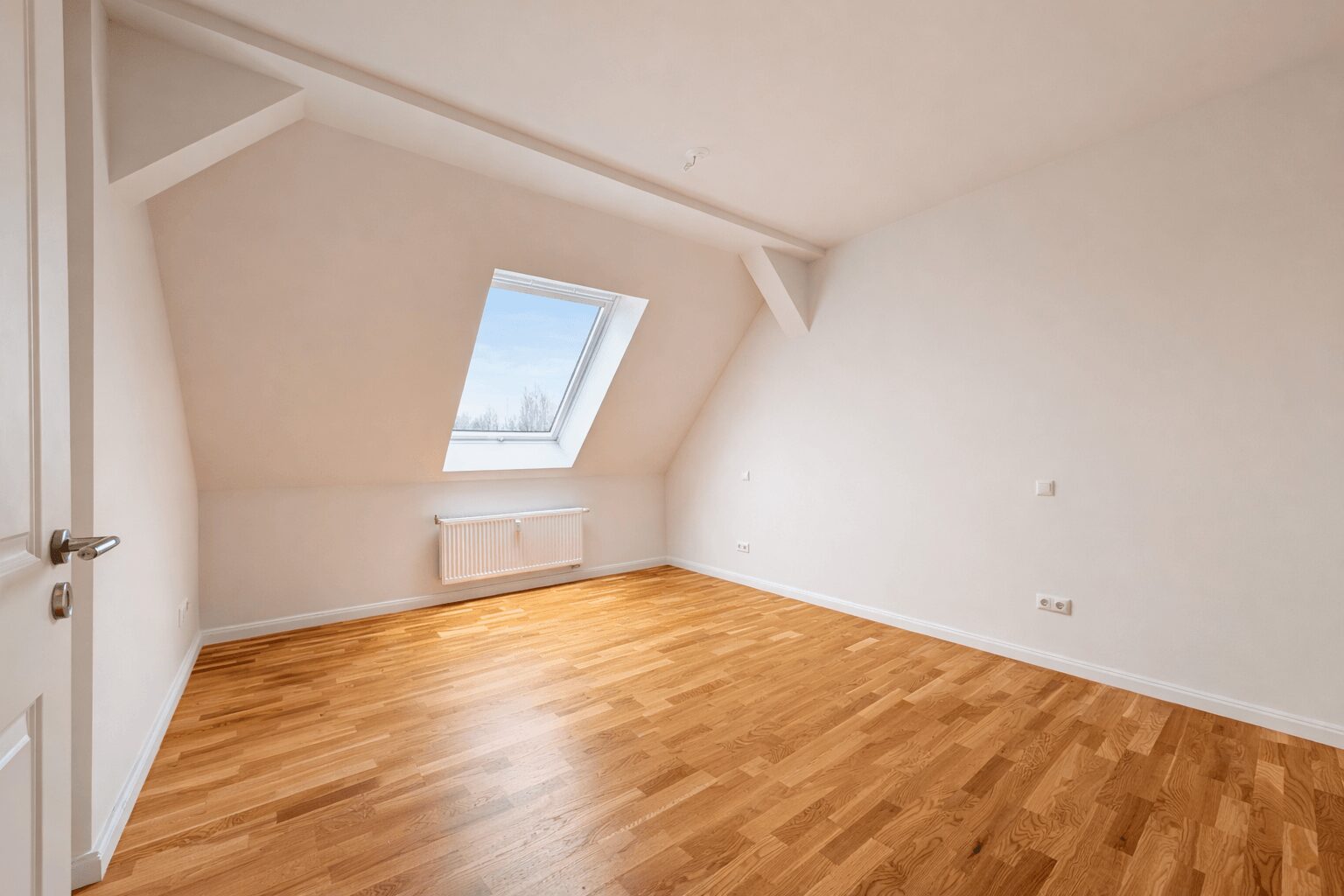 Empty attic room with sloped ceiling, a skylight window, and a wooden floor; radiator under the window.