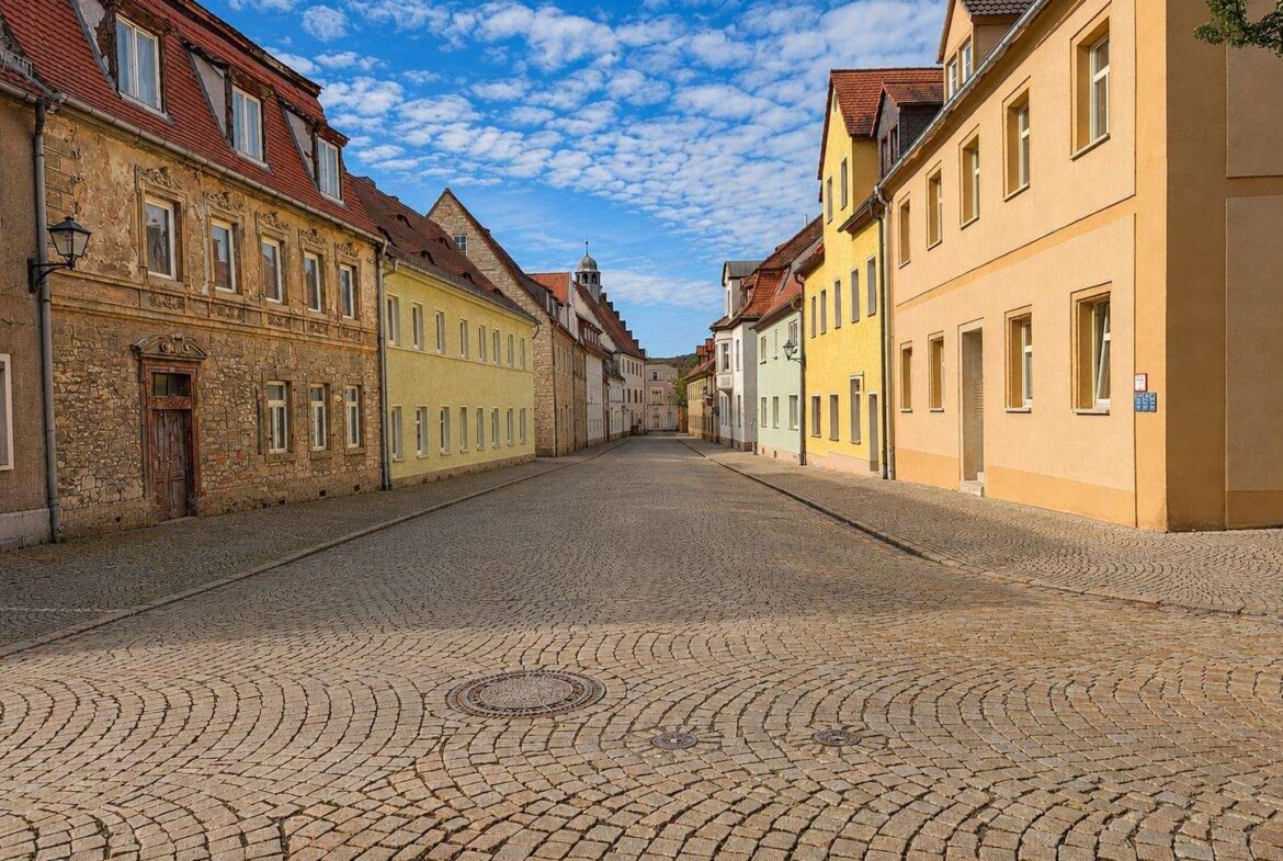 Sunlit cobblestone street lined with pastel houses in a quiet European town.