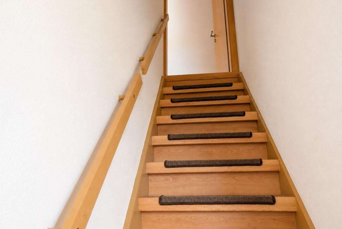 View up a narrow wooden staircase with a left handrail and carpeted treads toward a door at the top of the stairs.