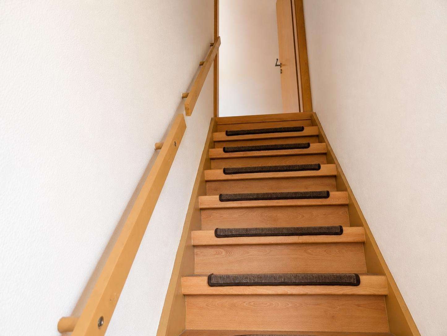 View up a narrow wooden staircase with a left handrail and carpeted treads toward a door at the top of the stairs.