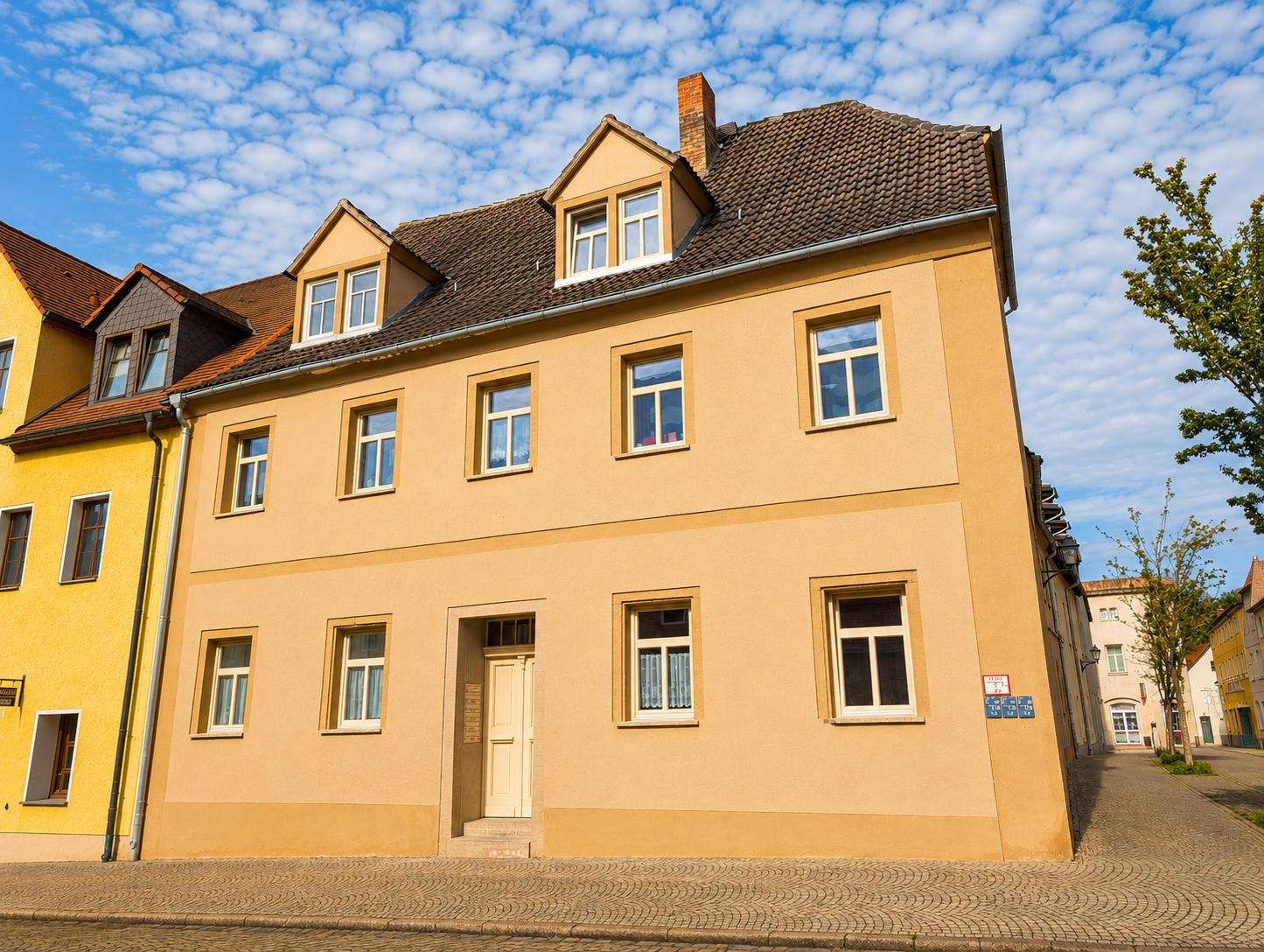 Peach-colored, three-story townhouse with white-framed windows on a cobblestone street under a blue, partly cloudy sky (with a yellow building on the left).