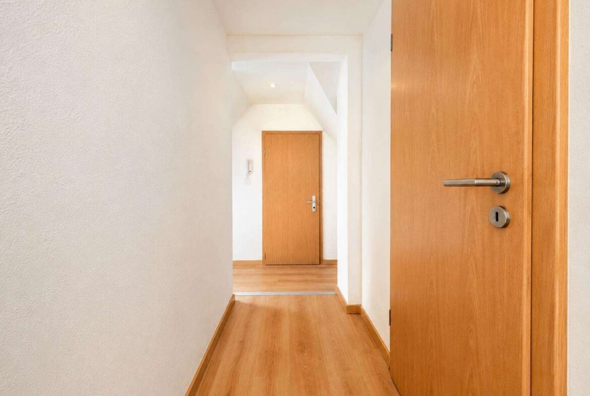 Narrow white hallway with light wood flooring and multiple wooden doors, viewer looking toward a closed door at the end.