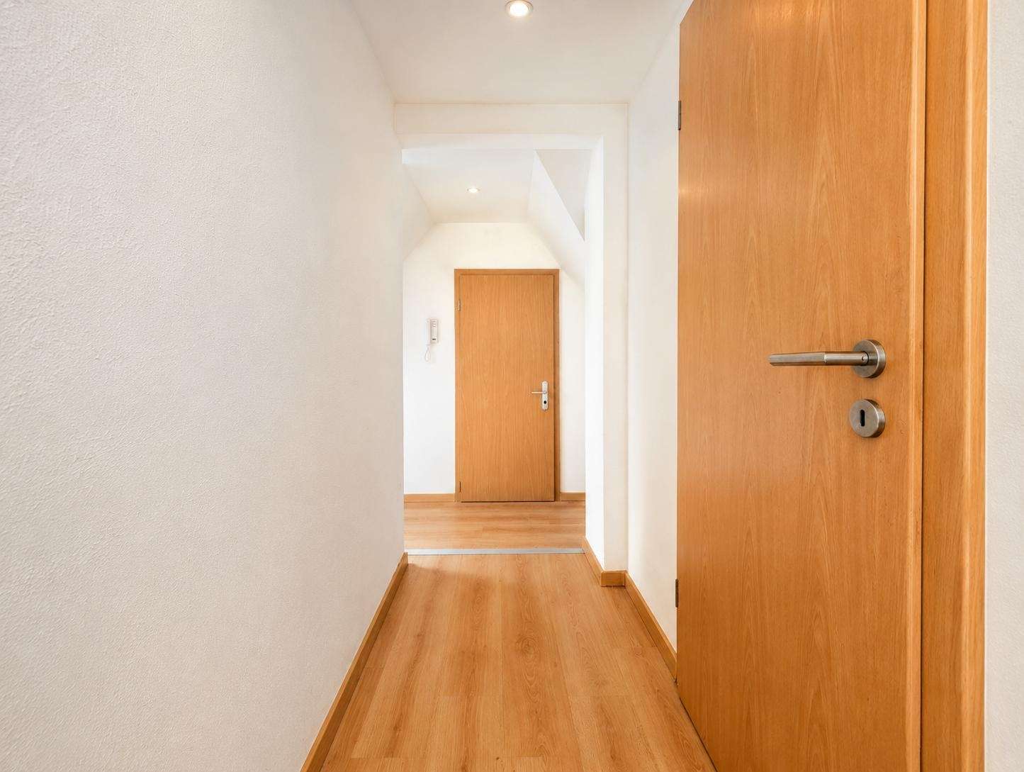 Narrow white hallway with light wood flooring and multiple wooden doors, viewer looking toward a closed door at the end.