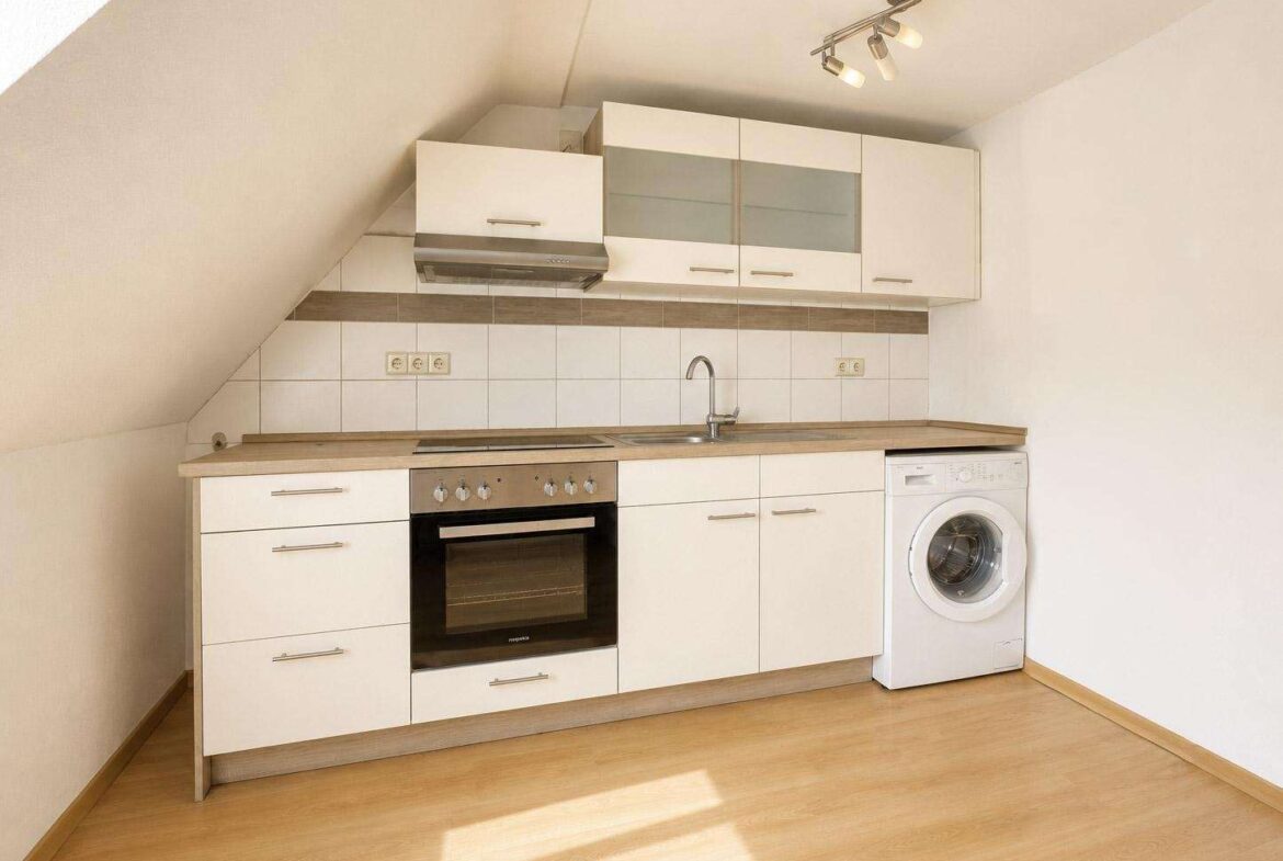 Compact white kitchen under a slanted ceiling with an oven, stove, range hood, sink, and a washing machine.