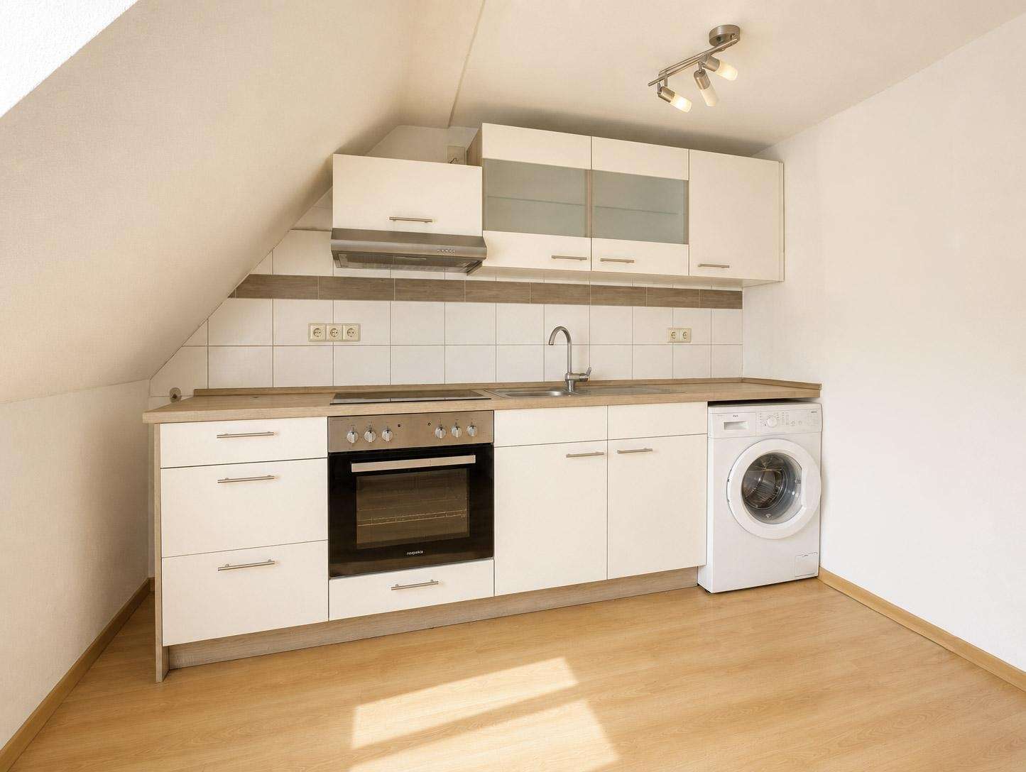 Compact white kitchen under a slanted ceiling with an oven, stove, range hood, sink, and a washing machine.