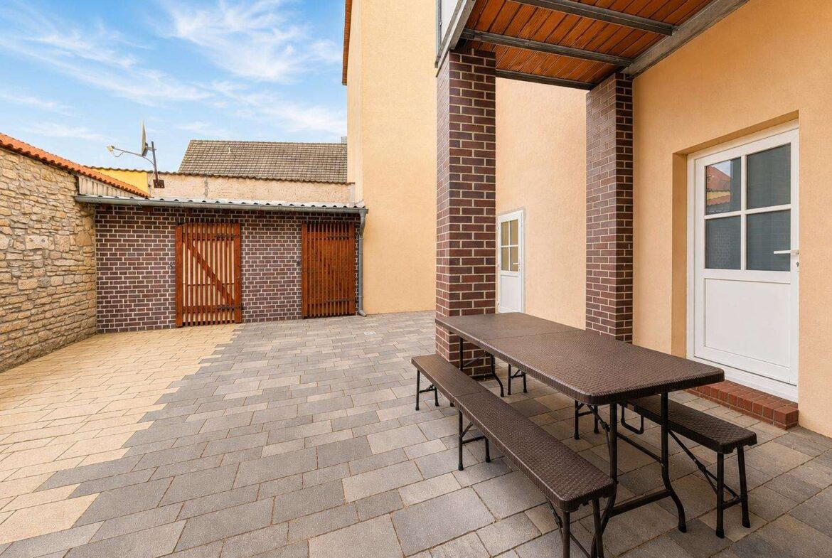 Outdoor paved courtyard with brick columns, a brown picnic table and benches, beige building walls, and wooden garage doors in the background.