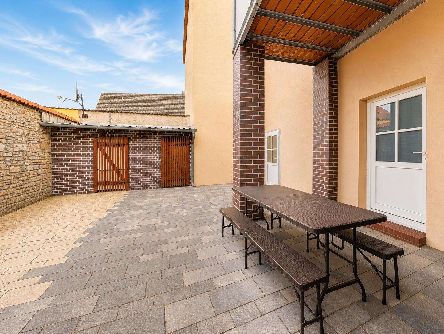 Outdoor paved courtyard with brick columns, a brown picnic table and benches, beige building walls, and wooden garage doors in the background.
