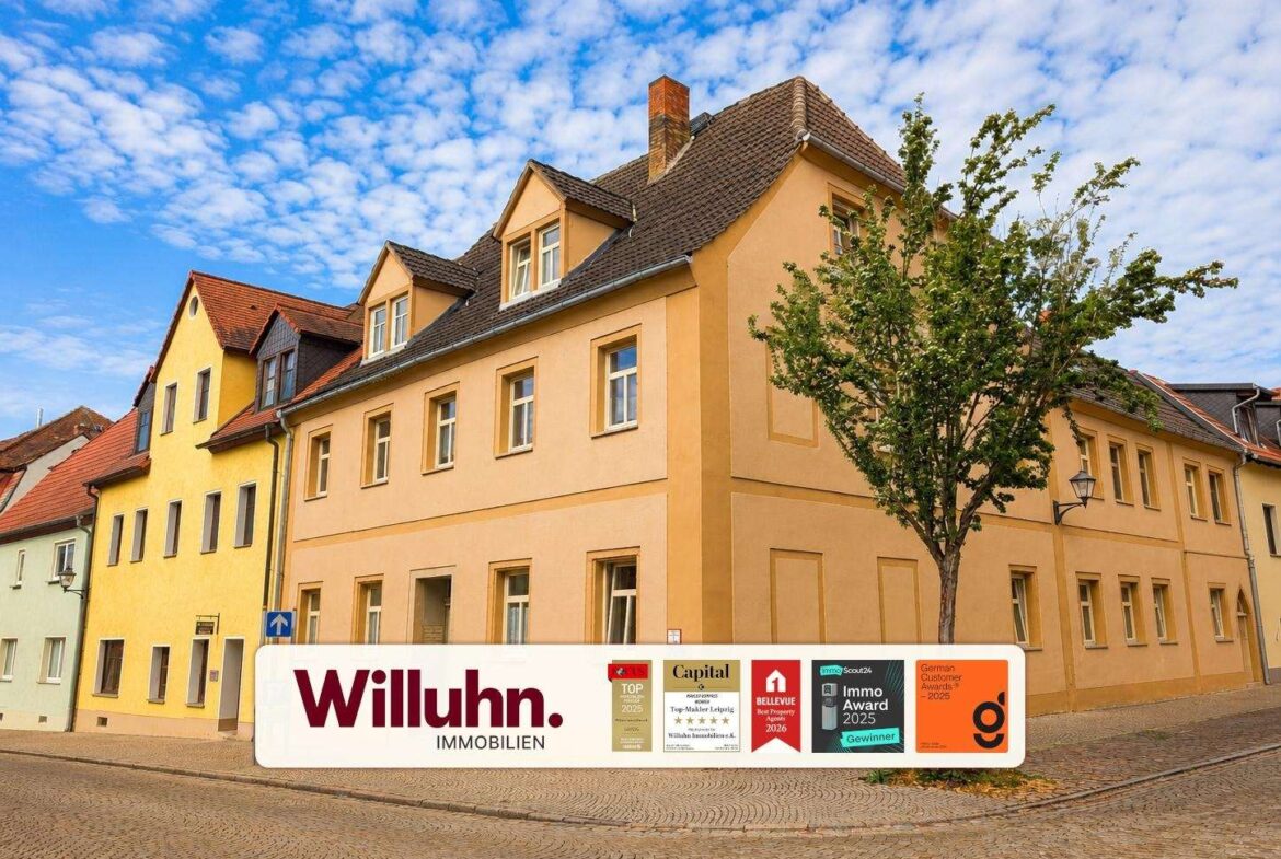 Pastel townhouses on a cobblestone street with a large Willuhn Immobilien banner in front of peach-colored buildings and a tree nearby.