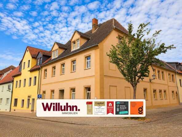 Pastel townhouses on a cobblestone street with a large Willuhn Immobilien banner in front of peach-colored buildings and a tree nearby.