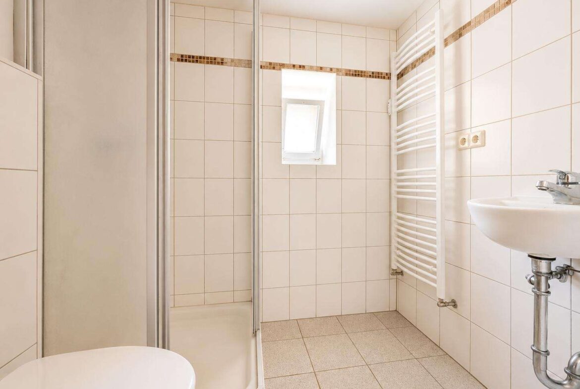 White tiled bathroom with a corner shower enclosure, wall-mounted sink, and a vertical white towel radiator.