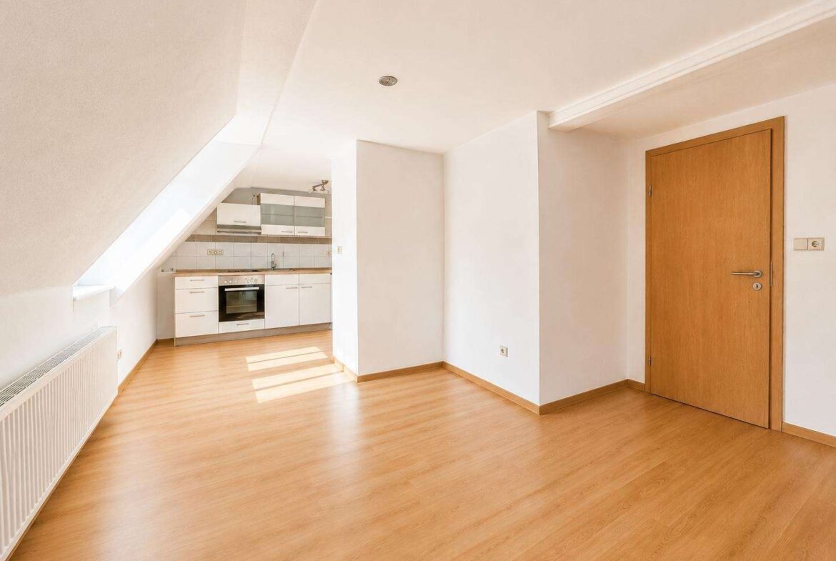 Bright, empty attic studio with sloped left ceiling, wooden floor, and a compact white kitchen at the back; door on the right bathed in sunlight.