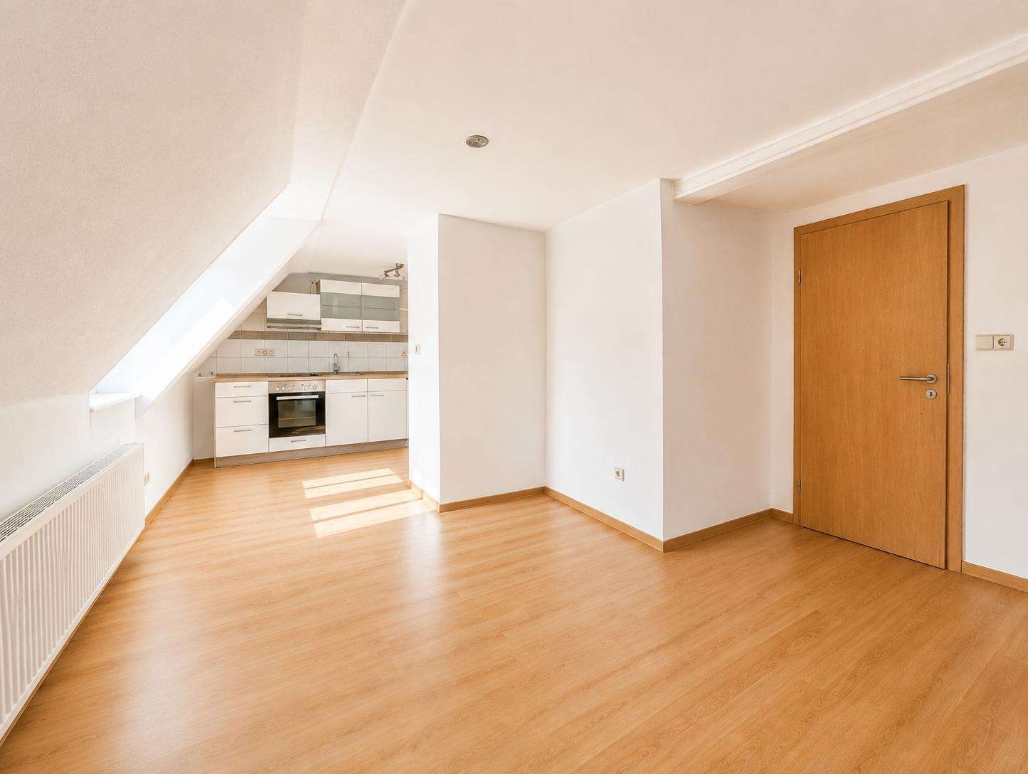 Bright, empty attic studio with sloped left ceiling, wooden floor, and a compact white kitchen at the back; door on the right bathed in sunlight.