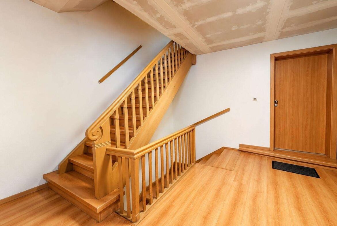 Wooden staircase with decorative curved newel post, light wood railing, and matching floor in a hallway near a closed wood door (under construction ceiling).