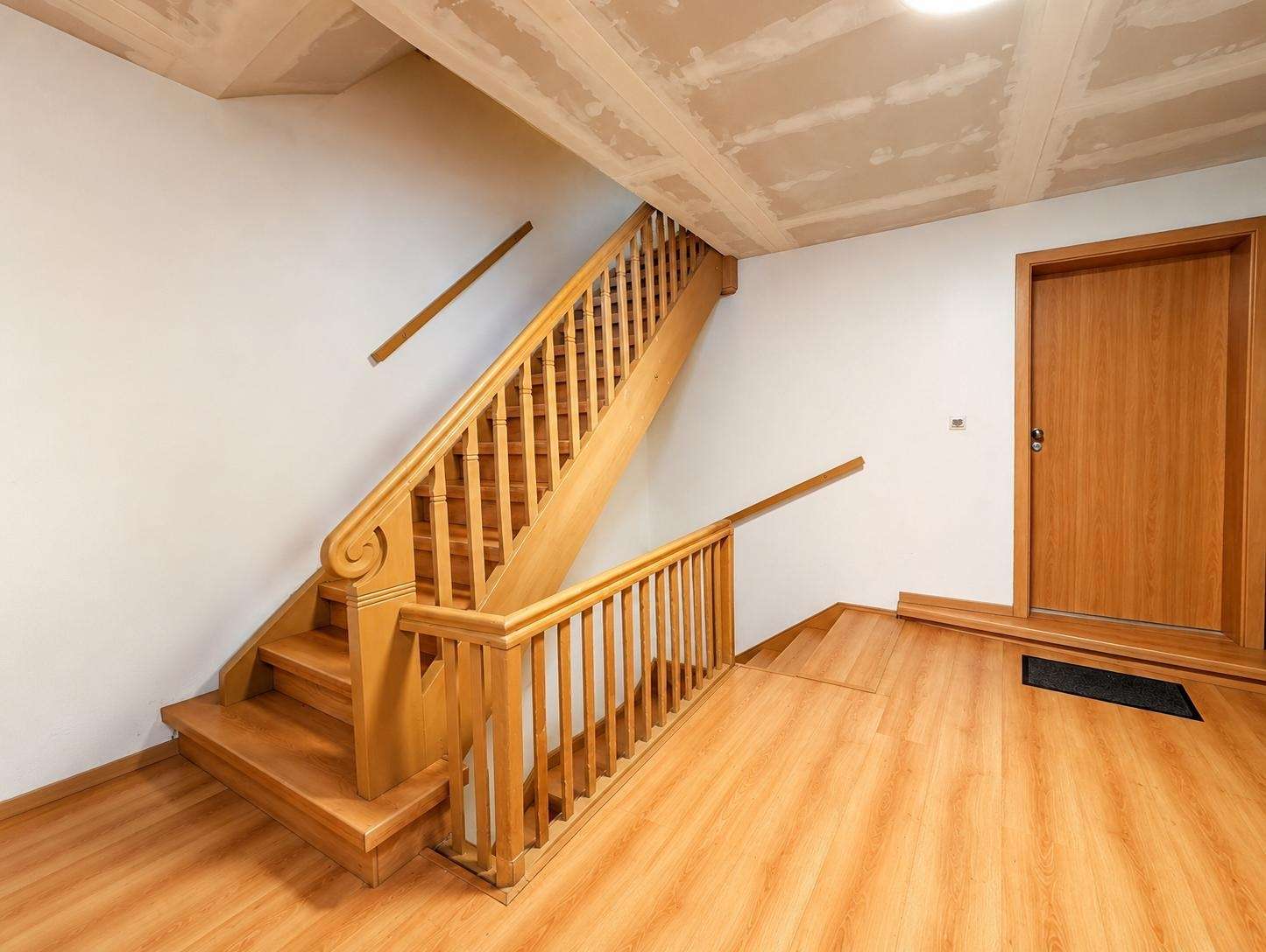 Wooden staircase with decorative curved newel post, light wood railing, and matching floor in a hallway near a closed wood door (under construction ceiling).