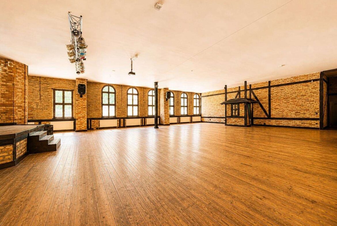 Spacious brick-walled rehearsal hall with a wooden floor, arched windows along one wall, and a small stage on the left under overhead lighting rig.