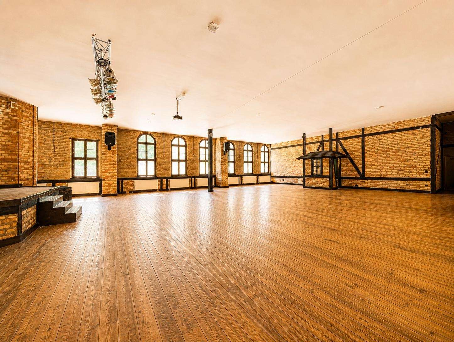 Spacious brick-walled rehearsal hall with a wooden floor, arched windows along one wall, and a small stage on the left under overhead lighting rig.
