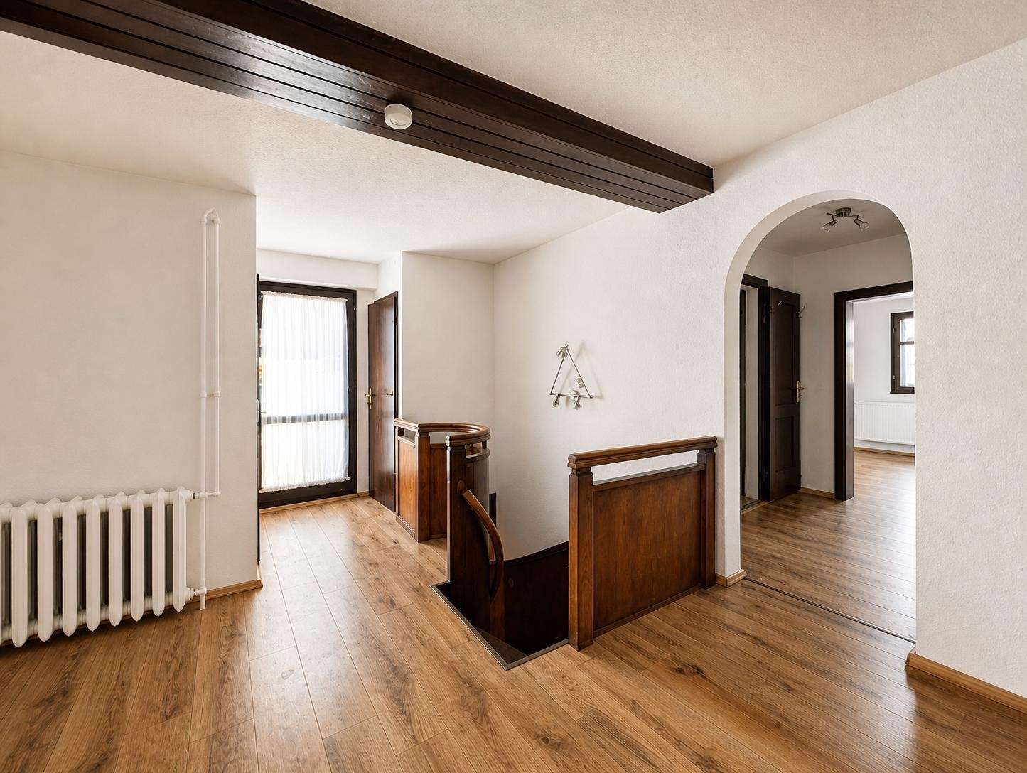 Bright hallway with a dark wooden ceiling beam, a wooden banister staircase, an arched doorway, and a radiator along the left wall.