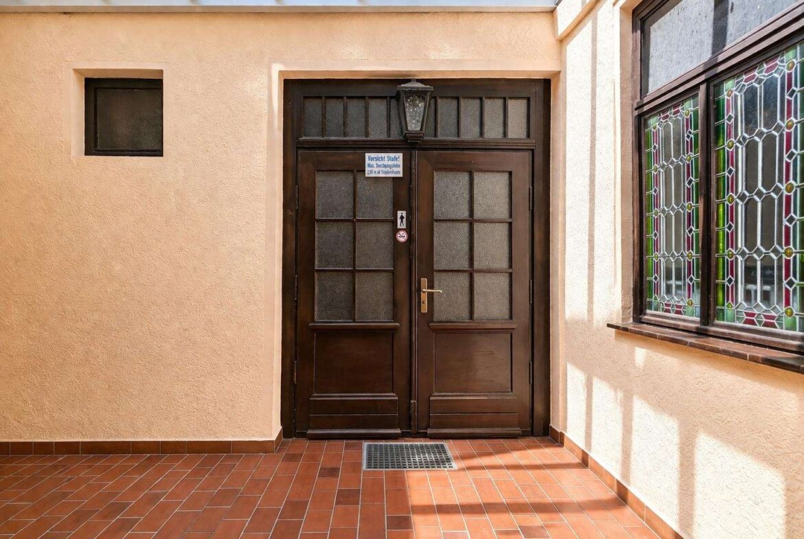 Double dark wooden doors with frosted glass panels, peach wall, lantern above, and a stained glass window to the right.