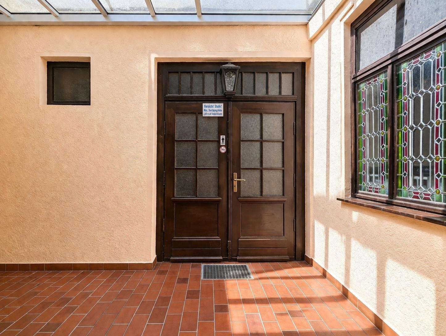 Double dark wooden doors with frosted glass panels, peach wall, lantern above, and a stained glass window to the right.