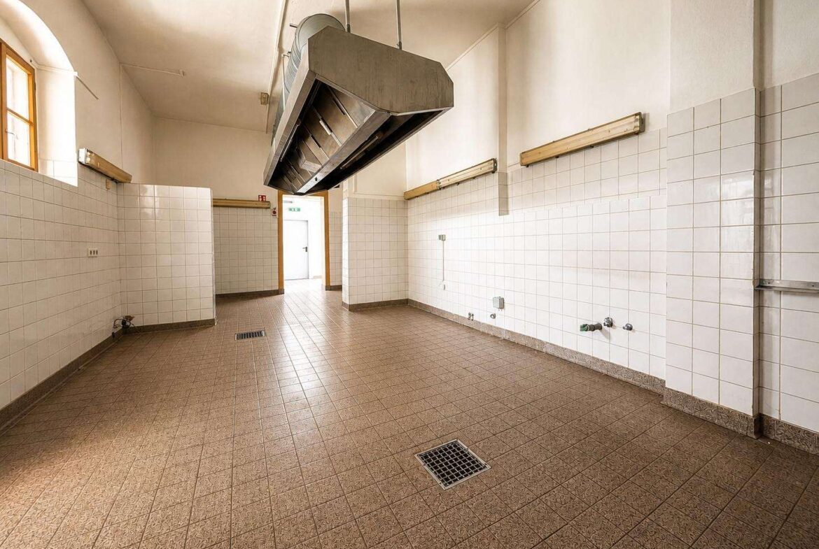 Empty industrial kitchen with a large ceiling-mounted exhaust hood over a tiled floor, bright and vacant