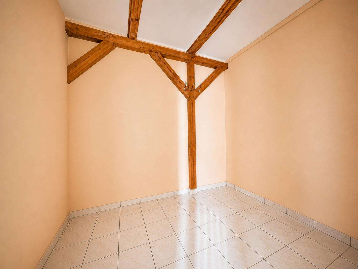 Empty corner room with peach walls, white tiled floor, and exposed wooden beams forming a triangular support in the center of the space.