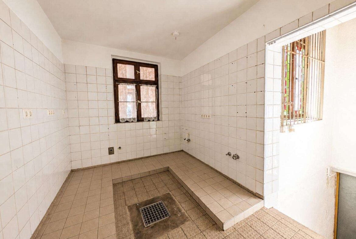 Empty tiled utility room with a central floor drain and a window with lace curtains on the wall.