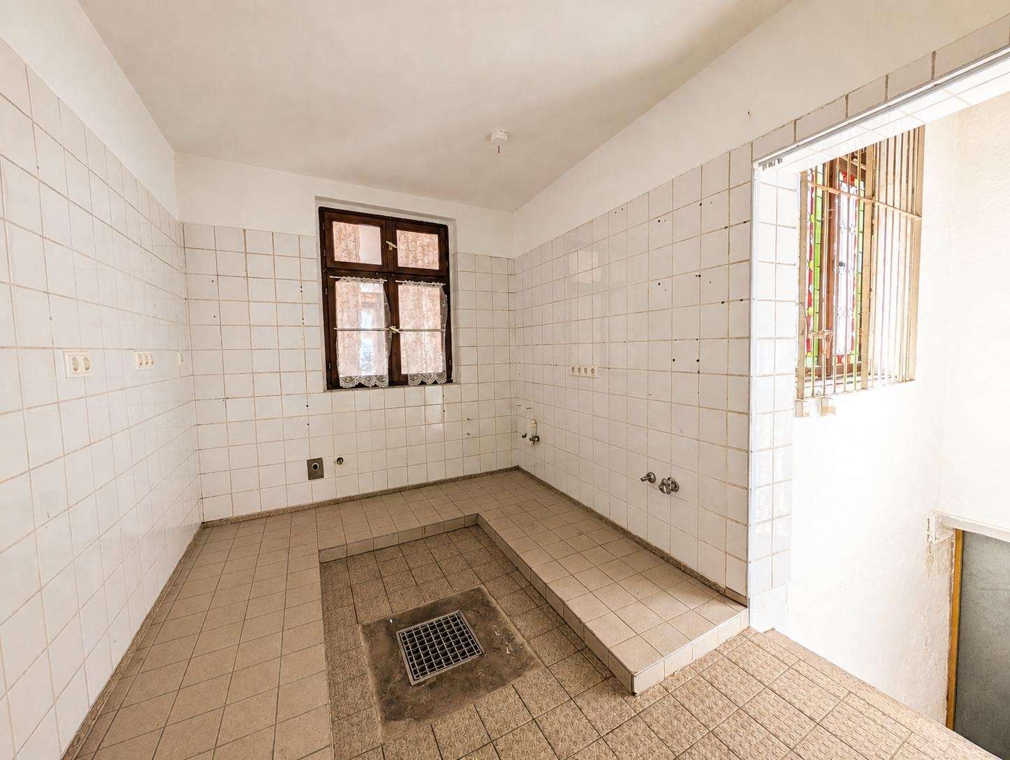 Empty tiled utility room with a central floor drain and a window with lace curtains on the wall.
