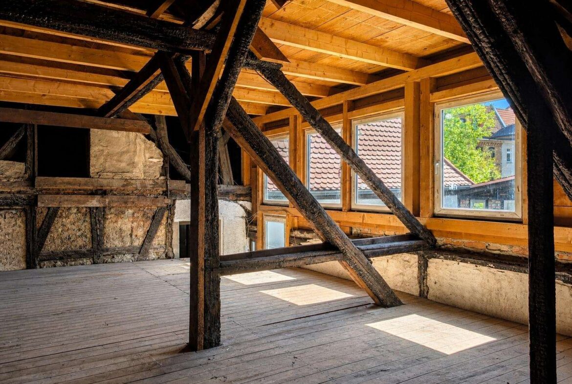 Sunlit attic with dark timber beams and diagonal supports, a wooden floor, and three windows looking out at red-tiled rooftops.