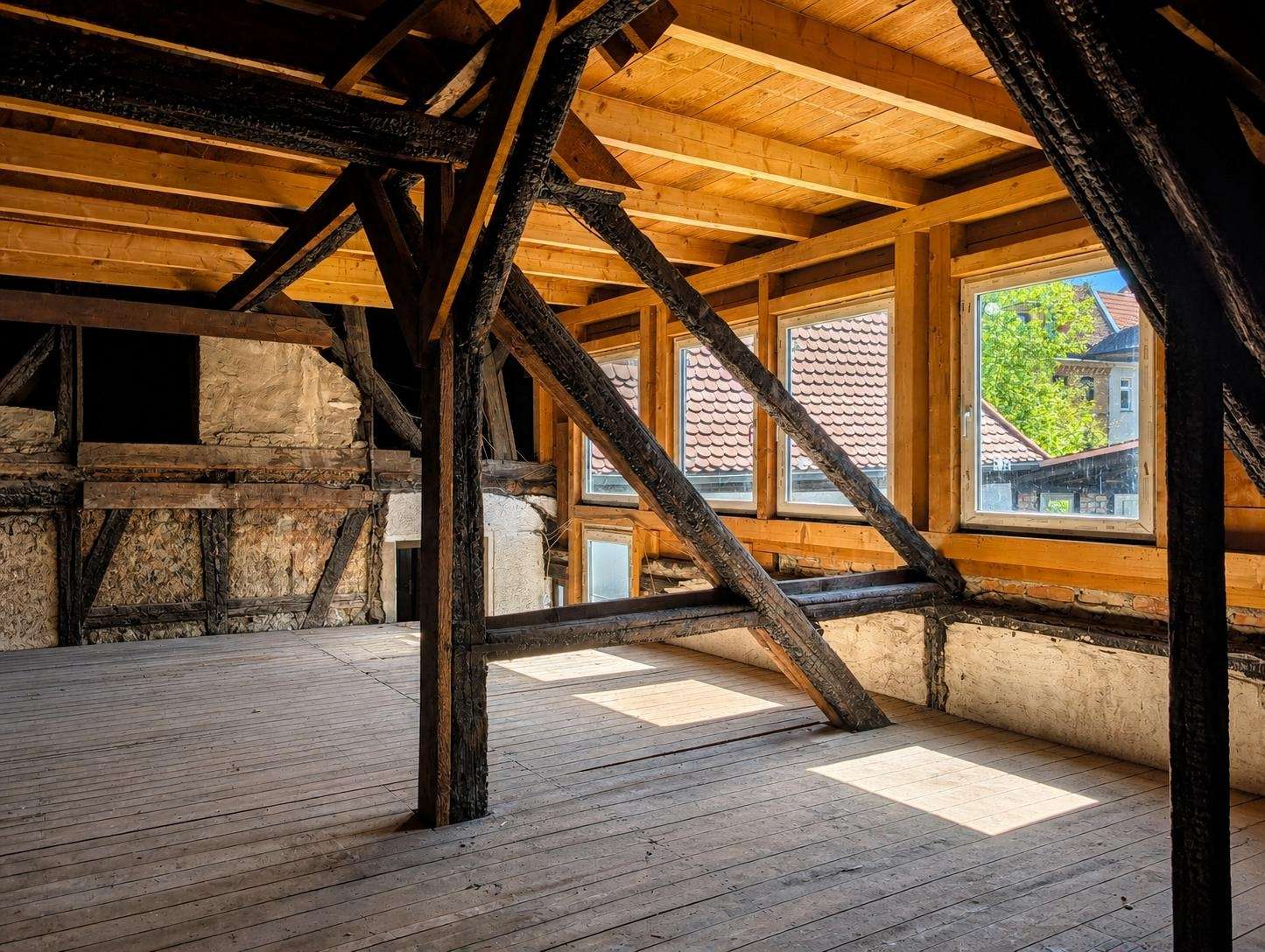 Sunlit attic with dark timber beams and diagonal supports, a wooden floor, and three windows looking out at red-tiled rooftops.