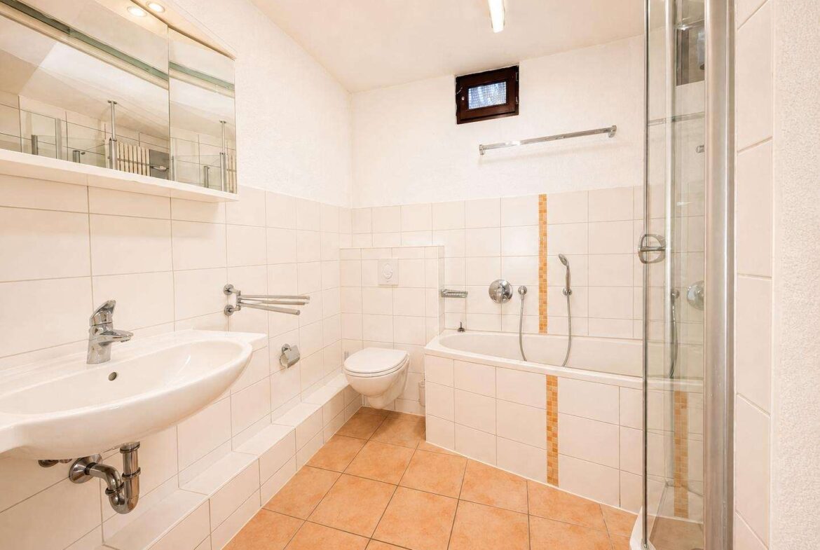 Bright bathroom with white tiled walls, wall-mounted sink, toilet, bathtub with handheld shower, and a glass shower enclosure with orange tile accents on the tub.