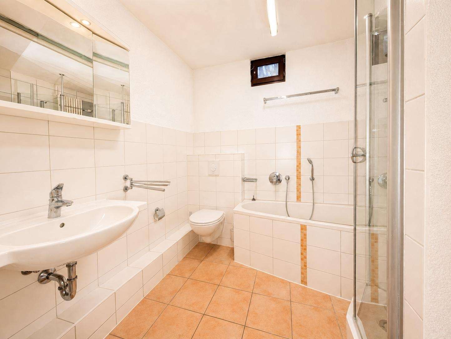 Bright bathroom with white tiled walls, wall-mounted sink, toilet, bathtub with handheld shower, and a glass shower enclosure with orange tile accents on the tub.