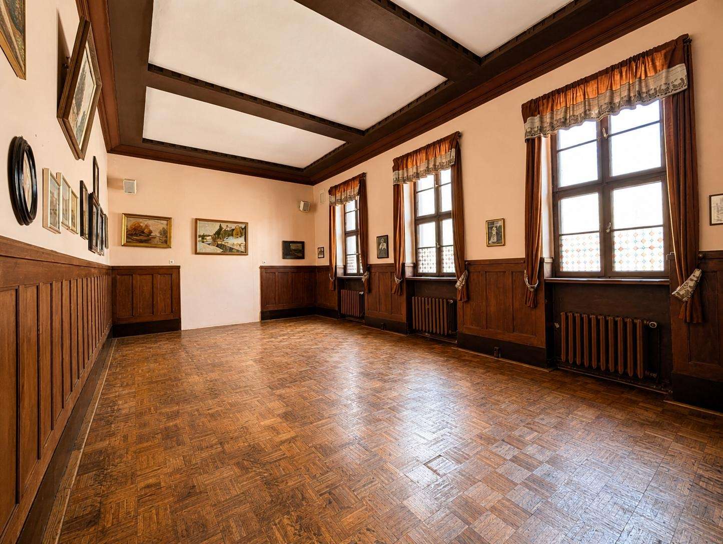 Sunlit, vintage hall with parquet floor, dark wood wainscoting, and tall windows with brown curtains and signage-like art on walls (interior gallery).