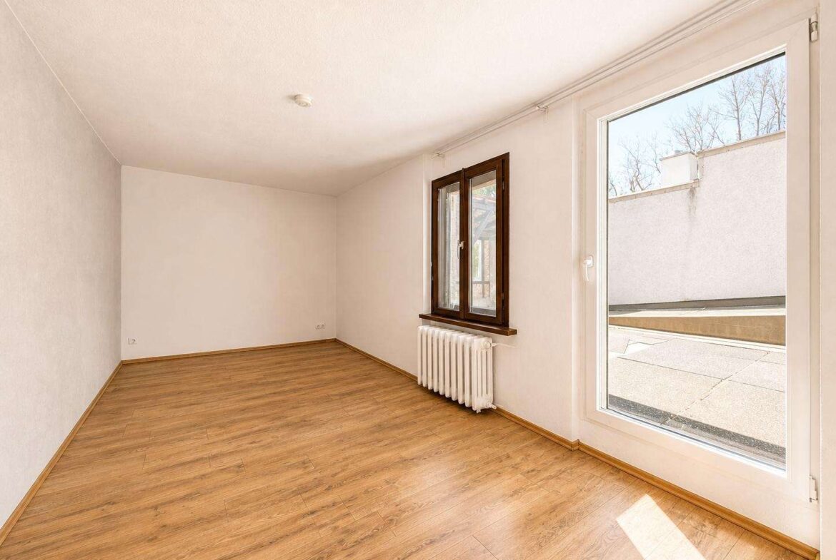Empty room with light wood flooring, white walls, a brown-framed window, and a glass door opening to an outdoor terrace; radiator beneath the window.