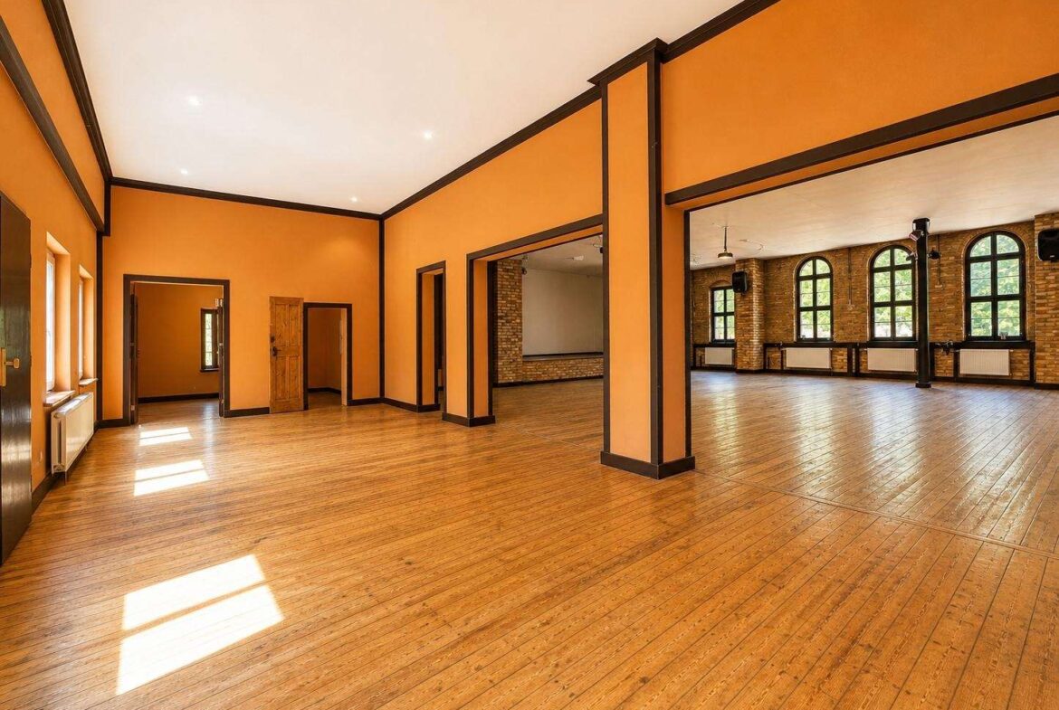 Sunlit, empty event hall with orange walls, dark trim, and a wooden floor; doorways lead to smaller rooms and arched windows in the back area.
