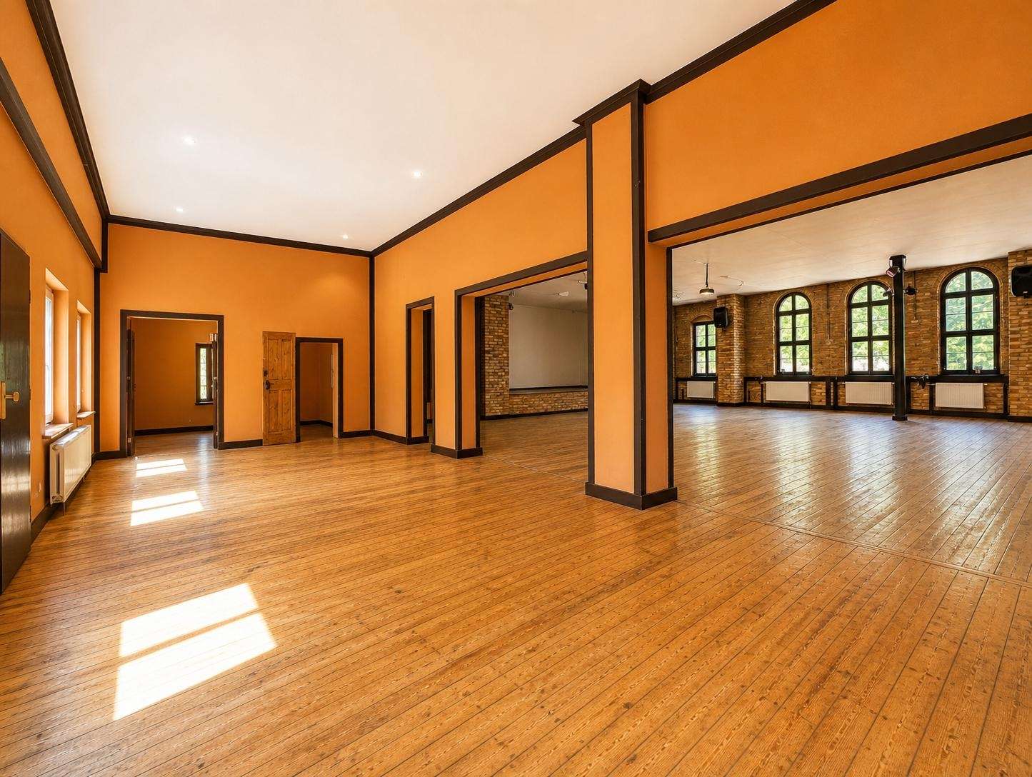 Sunlit, empty event hall with orange walls, dark trim, and a wooden floor; doorways lead to smaller rooms and arched windows in the back area.